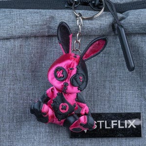 May include: A pink and black bunny keyring with a stitched design. The keyring is attached to a silver ring and chain. The bunny has a unique, stylized appearance. A black rectangular tag with the text "STLFLIX" is also visible.