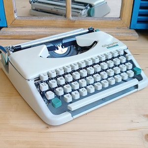 May include: A cream-colored Olympia Traveller typewriter featuring a QWERTZ keyboard layout with white keys, teal accent keys, and a metal carriage. The brand name Olympia Traveller is printed in teal on the top right corner of the machine casing.