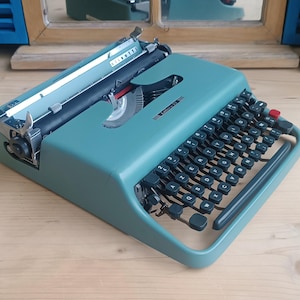 May include: A teal Olivetti Lettera 22 typewriter featuring a QWERTY keyboard layout with black circular keys, a metal carriage, and a manual return lever. The device has a sleek, low-profile design with a metallic finish and a red key on the right.