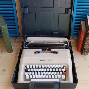 May include: A beige Olivetti Lettera 35 typewriter in a black case. The typewriter features white keys with black lettering and a red return key. The case is open, displaying the typewriter's internal mechanisms.