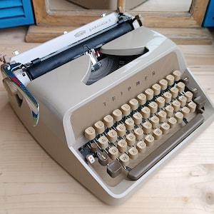 Peut inclure: Machine à écrire Triumph Galaxie-F vintage, beige et marron. Le clavier complet comporte des touches rondes blanc cassé. Le nom de la marque est visible. Design rétro.