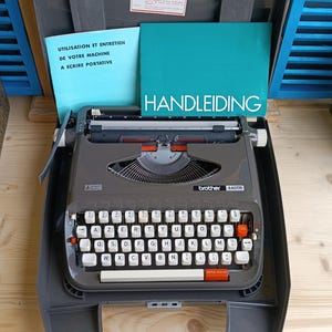 May include: A gray Brother 440TR typewriter with white keys and a red tab key. The typewriter has a silver carriage and a paper guide. The image includes manuals in French and Dutch.