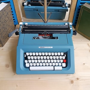 Può includere: Una macchina da scrivere Olivetti Studio 46 turchese vintage con tasti bianchi e un tasto di ritorno rosso. La macchina da scrivere è posizionata su una superficie di legno chiaro, con uno specchio e libri sullo sfondo. Il nome del modello è visibile.
