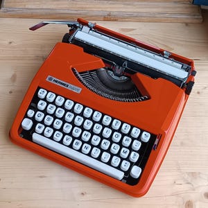 Pode incluir: Uma máquina de escrever Hermes Baby laranja com teclas brancas e letras pretas. A máquina de escrever tem uma alavanca de retorno de carro prateada. O nome da marca é visível na frente. Disposição das teclas QWERTY.