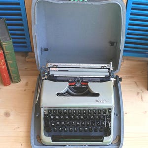 May include: Vintage Olympia typewriter in a gray case. The typewriter is light green with black keys and a red ribbon. The case is open, revealing the typewriter inside. The keys are arranged in a QWERTY layout.