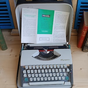May include: A vintage Olympia De Luxe typewriter in its black carrying case. The typewriter is light grey with white keys and teal accents. The case is open, revealing the machine and instruction manual. The Olympia logo is visible.