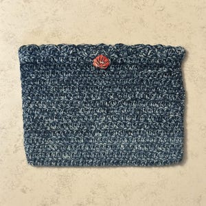 May include: A rectangular crochet pouch crafted from variegated dark blue and grey yarn. The top edge features a decorative scalloped border and a single, centered, orange-red shell-shaped button closure, providing a secure and textured finish.