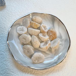 May include: A decorative, scalloped-edge ceramic dish with a silver rim, filled with small, white and beige geode specimens. The geodes are cut open, revealing crystal formations inside. The dish has a marbled, gray and white pattern.