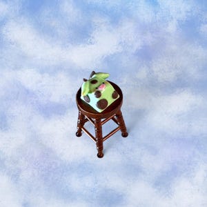 Può includere: Un oggetto stravagante a forma di mucca, verde chiaro con macchie marroni, poggia su uno sgabello di legno scuro. Lo sgabello ha una seduta rotonda e quattro gambe. La mucca ha un naso rosa e orecchie verdi.