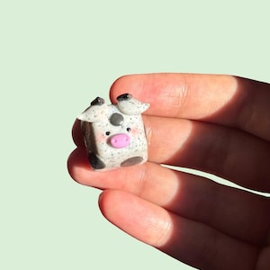 Puede incluir: Pequeña figura de vaca hecha a mano con cuerpo blanco y gris moteado. La vaca tiene orejas negras, una mancha negra y una nariz rosa. La figura se sostiene en una mano sobre un fondo verde claro.