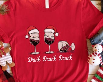 May include: A red t-shirt with a graphic of three wine glasses, two upright and one tipped over, each wearing a Santa hat. The text "Drink Drank Drunk" is below the glasses. Christmas decorations surround the shirt.