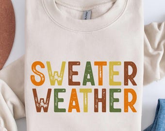 May include: A cream-colored sweatshirt with the words "SWEATER WEATHER" in orange, green, yellow, and brown block letters. The sweatshirt is folded with a brown hat and blue jeans in the background.