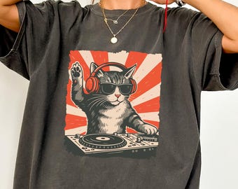 May include: Dark grey t-shirt featuring a graphic of a cat DJ wearing sunglasses and headphones, with a red and white sunburst background. The cat is positioned behind a turntable, with one paw raised.