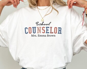 May include: White t-shirt with the words "School COUNSELOR" in blue and red lettering. Below the text is "Mrs. Emma Brown" in red. The shirt is a casual style, suitable for everyday wear.