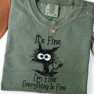 May include: A moss green t-shirt with the phrase "It's Fine, I'm Fine, Everything Is Fine" and a cartoon cat graphic. A gold necklace with a pendant is draped across the shirt. A brown hat and dried plant are in the background.
