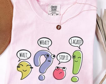 May include: A light pink Comfort Colors t-shirt with a crew neck, featuring cartoon speech bubbles with the words "WAIT," "WHAT?," "STOP IT," and "I AGREE!" A gold necklace with a pendant is draped on the shirt.