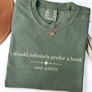 Op de afbeelding: Mosgroen t-shirt met de quote "I should infinitely prefer a book." van Jane Austen. Een gouden ketting met een hanger ligt op het shirt. Een bruine hoed en gedroogde palmbladeren zijn op de achtergrond.