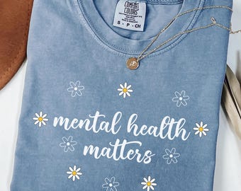 Könnte beinhalten: Eisblaues T-Shirt mit dem weißen Schriftzug "mental health matters", verziert mit Gänseblümchen-Grafiken. Eine goldene Halskette mit Anhänger liegt auf dem Shirt. Das Shirt ist von der Marke Comfort Colors, Größe S-P-CH.