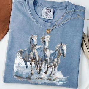 May include: Ice blue Comfort Colors t-shirt featuring a graphic of three white horses running through water. The shirt has a crew neck and short sleeves. A gold necklace with a pendant is draped across the shirt.