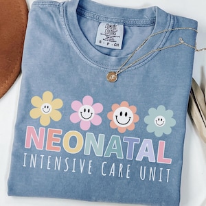 May include: Ice blue t-shirt with a floral design and the words "Neonatal Intensive Care Unit" in pastel colors. The shirt has a crew neck and short sleeves. A gold necklace with a pendant is draped across the shirt.