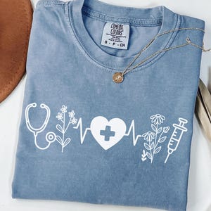 May include: Ice blue t-shirt with a white graphic of a stethoscope, flowers, a heartbeat line, a heart with a cross, and a syringe. The shirt has a crew neck and short sleeves. A gold necklace is draped across the shirt.