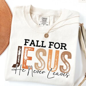 K&ouml;nnte beinhalten: Elfenbeinfarbenes Comfort Colors T-Shirt mit dem Text "FALL FOR JESUS He Never Leaves" in Braun, Orange und Beige. Eine goldene Halskette und ein brauner Hut sind im Hintergrund zu sehen. Das Shirt hat einen Rundhalsausschnitt.