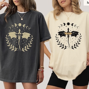 May include: Two women wearing oversized t-shirts with a graphic print of a dragon with wings spread, surrounded by a circle of moons and stars. The t-shirt on the left is dark gray and the t-shirt on the right is ivory.
