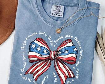 May include: A light blue t-shirt with a red, white, and blue bow with stars. The bow is surrounded by the text "America Land of The free * America land of The free * America land of The free * America land of The free".