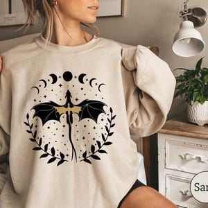 May include: A beige sweatshirt with a black and gold dragon design. The dragon is surrounded by a circle of crescent moons and stars. The text "Sand" is visible in the lower right corner of the image.