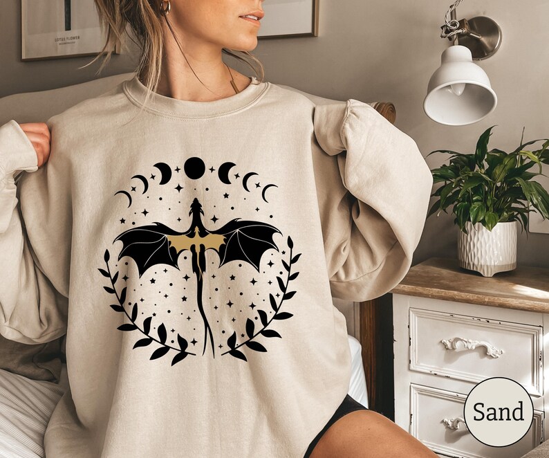 May include: A beige sweatshirt with a black and gold dragon design. The dragon is surrounded by a circle of crescent moons and stars. The text "Sand" is visible in the lower right corner of the image.