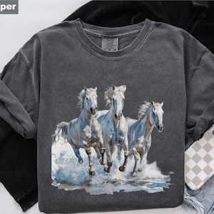 May include: A dark gray t-shirt featuring a graphic of three white horses running through water. The horses have flowing manes and tails, and the water is depicted with blue and white splashes. The shirt has a crew neck and short sleeves.