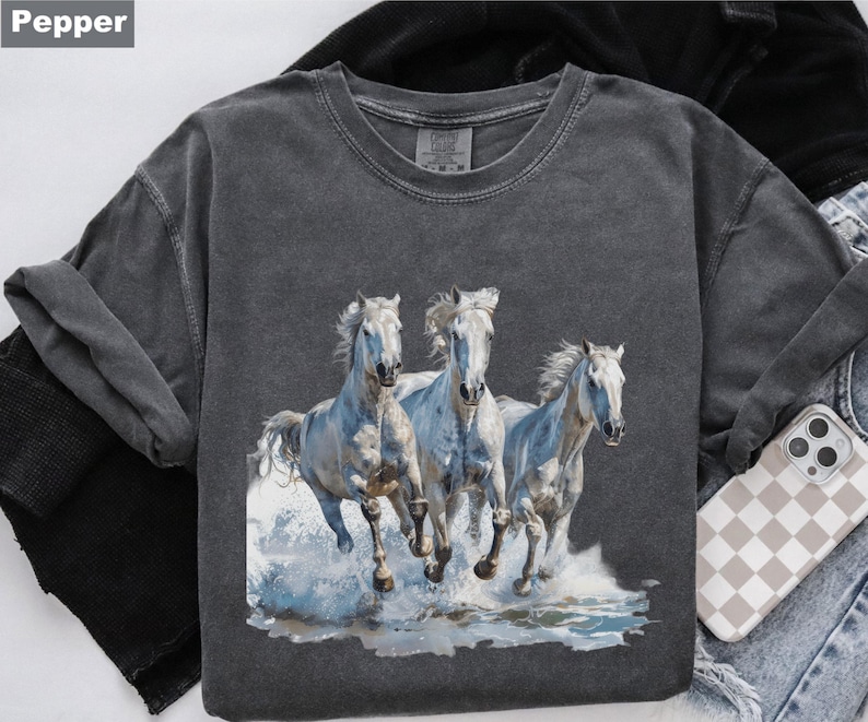 May include: A dark gray t-shirt featuring a graphic of three white horses running through water. The horses have flowing manes and tails, and the water is depicted with blue and white splashes. The shirt has a crew neck and short sleeves.