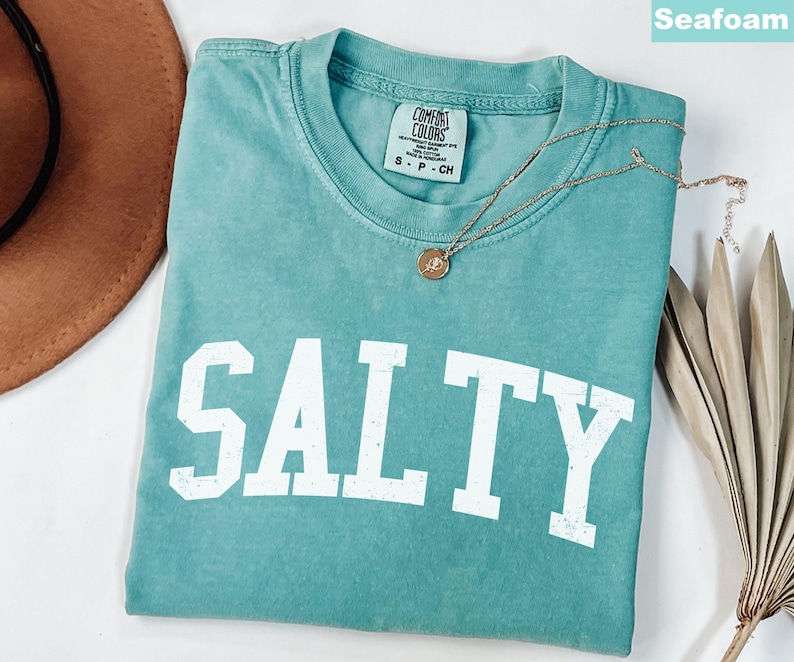 Puede incluir: Camiseta verde agua con la palabra "SALTY" impresa en blanco en la parte delantera. La camiseta est&aacute; hecha de una tela suave y c&oacute;moda y tiene un corte holgado.