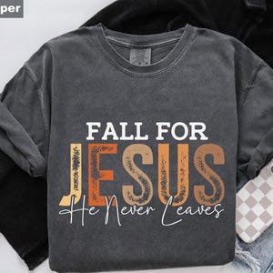 K&ouml;nnte beinhalten: Ein dunkelgraues T-Shirt mit dem Schriftzug "FALL FOR JESUS He Never Leaves" in Wei&szlig;, Orange und Braun. Das T-Shirt ist gefaltet, im Hintergrund ist eine Handyh&uuml;lle zu sehen.