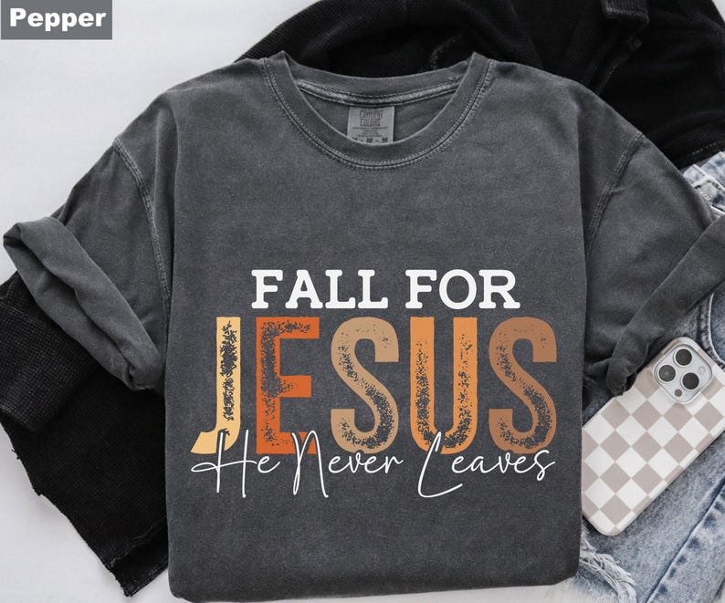 K&ouml;nnte beinhalten: Ein dunkelgraues T-Shirt mit dem Schriftzug "FALL FOR JESUS He Never Leaves" in Wei&szlig;, Orange und Braun. Das T-Shirt ist gefaltet, im Hintergrund ist eine Handyh&uuml;lle zu sehen.