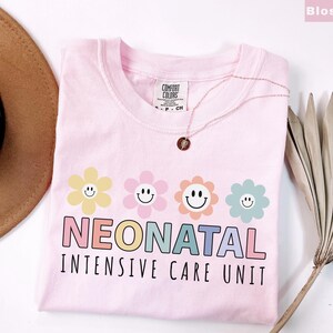 May include: A light pink t-shirt with the words "NEONATAL INTENSIVE CARE UNIT" in colorful letters. Above the text are four smiling flower graphics in yellow, pink, orange, and blue. A gold necklace rests on the shirt.