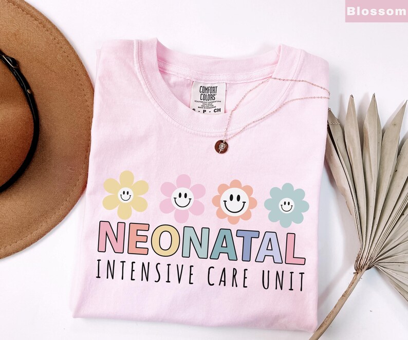 May include: A light pink t-shirt with the words "NEONATAL INTENSIVE CARE UNIT" in colorful letters. Above the text are four smiling flower graphics in yellow, pink, orange, and blue. A gold necklace rests on the shirt.