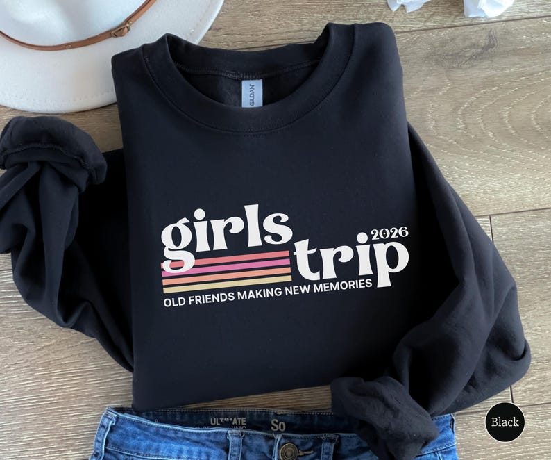 May include: Black sweatshirt with the words "girls trip 2026" in white, with a retro-style stripe design in pink, orange, and yellow. The text "OLD FRIENDS MAKING NEW MEMORIES" is below the main text. The sweatshirt is folded on top of blue jeans.