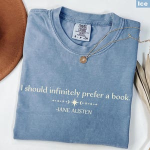 Op de afbeelding: IJsblauw Comfort Colors T-shirt met de quote "I should infinitely prefer a book." van Jane Austen. Een gouden ketting met hanger ligt op het shirt. Een bruine hoed en gedroogde bladeren zijn op de achtergrond.