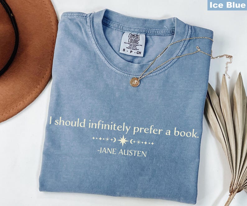 Op de afbeelding: IJsblauw Comfort Colors T-shirt met de quote "I should infinitely prefer a book." van Jane Austen. Een gouden ketting met hanger ligt op het shirt. Een bruine hoed en gedroogde bladeren zijn op de achtergrond.