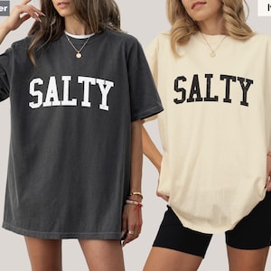 Puede incluir: Dos camisetas de gran tama&ntilde;o con la palabra "SALTY" impresa en una fuente grande y en negrita. La camiseta de la izquierda es de color gris oscuro con letras blancas. La camiseta de la derecha es de color marfil claro con letras negras.