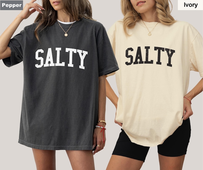 Puede incluir: Dos camisetas de gran tama&ntilde;o con la palabra "SALTY" impresa en una fuente grande y en negrita. La camiseta de la izquierda es de color gris oscuro con letras blancas. La camiseta de la derecha es de color marfil claro con letras negras.