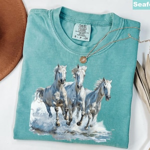 May include: Seafoam green t-shirt featuring a graphic of three white horses running through water. The shirt has a crew neck and short sleeves. A gold necklace with a pendant is draped across the shirt. The shirt is labeled "Comfort Colors".