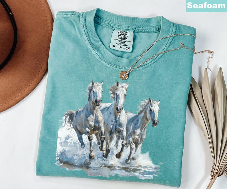 May include: Seafoam green t-shirt featuring a graphic of three white horses running through water. The shirt has a crew neck and short sleeves. A gold necklace with a pendant is draped across the shirt. The shirt is labeled "Comfort Colors".