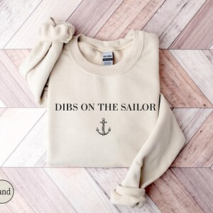 May include: Sand-colored sweatshirt with the text "DIBS ON THE SAILOR" and an anchor graphic. The crew neck sweatshirt has long sleeves with ribbed cuffs. The Gildan brand tag is visible on the inside of the collar.