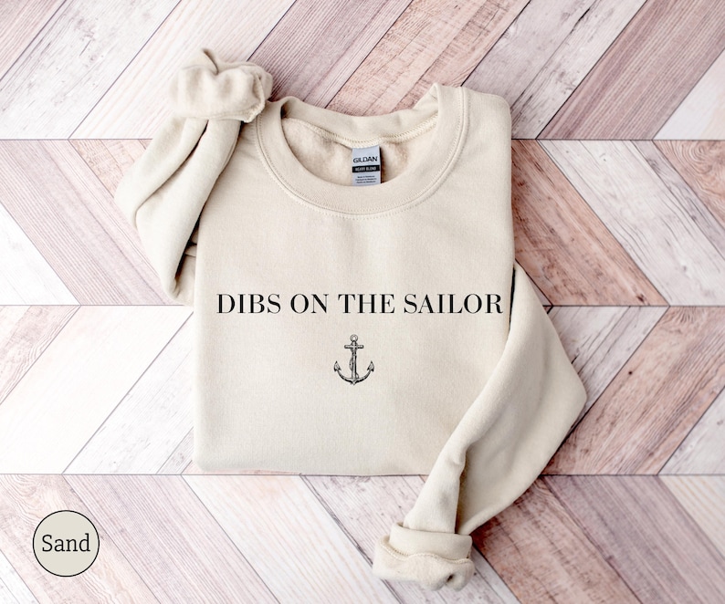 May include: Sand-colored sweatshirt with the text "DIBS ON THE SAILOR" and an anchor graphic. The crew neck sweatshirt has long sleeves with ribbed cuffs. The Gildan brand tag is visible on the inside of the collar.