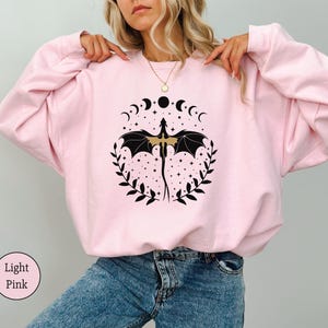 May include: A light pink sweatshirt with a black and gold dragon design. The dragon is surrounded by a circle of crescent moons and stars. The sweatshirt has a relaxed fit and is perfect for everyday wear.