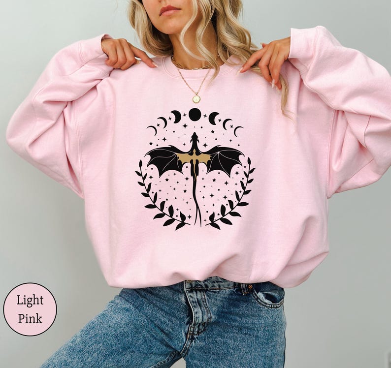 May include: A light pink sweatshirt with a black and gold dragon design. The dragon is surrounded by a circle of crescent moons and stars. The sweatshirt has a relaxed fit and is perfect for everyday wear.