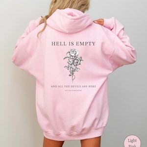May include: Light pink hoodie with a hood and long sleeves. The back features the text "HELL IS EMPTY" above a black and white rose and snake illustration. Below the illustration is the text "AND ALL THE DEVILS ARE HERE" and "WILLIAM SHAKESPEARE".