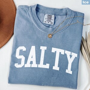 Puede incluir: Una camiseta azul claro con la palabra "SALTY" impresa en blanco en la parte delantera. La camiseta est&aacute; hecha de una tela suave y c&oacute;moda y tiene un corte holgado.